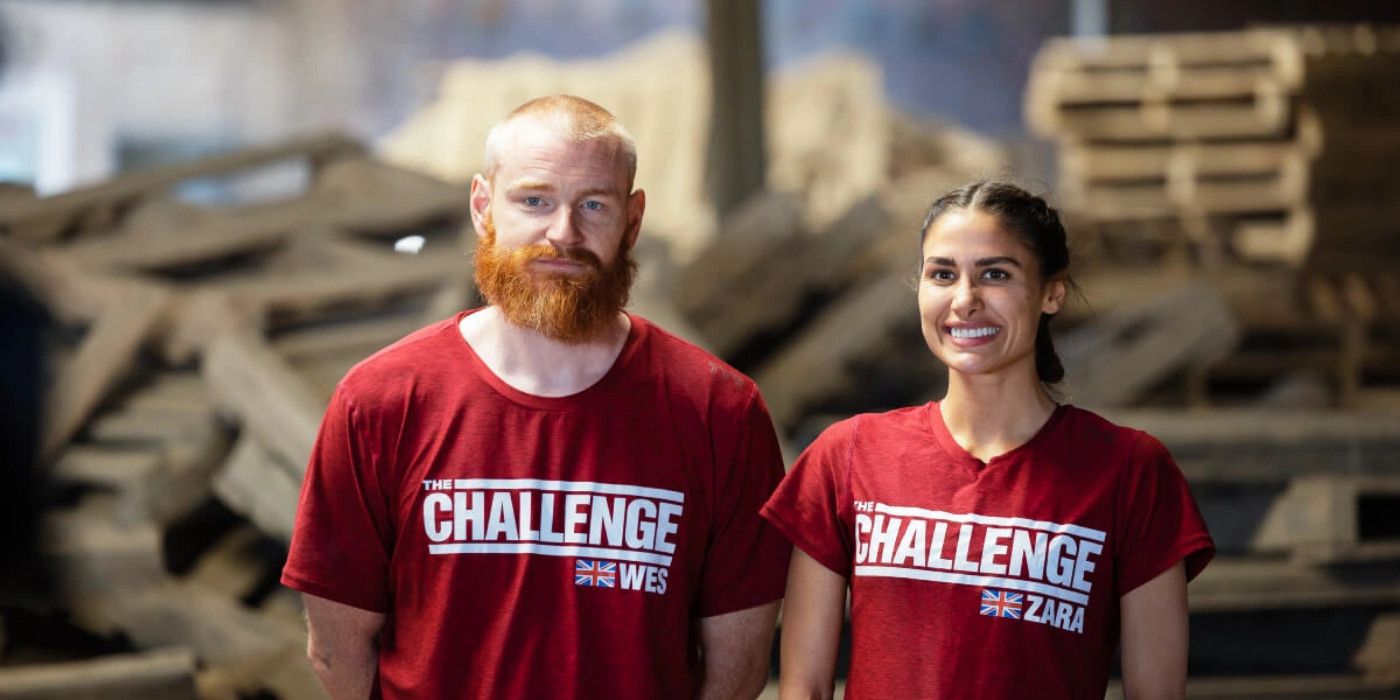 Zara Zoffany standing beside Wes Bergmann on The Challenge: World Championship. They are wearing red shirts with The Challenge logo and the UK flag. They are standing against a backdrop of debri in a warehouse.