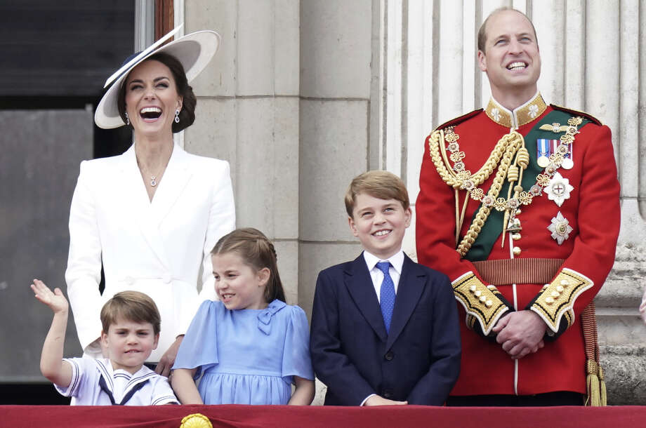 Catherine, la princesa de Gales, sonríe ampliamente con un traje blanco y un sombrero blanco de gran tamaño junto a su esposo, el príncipe William, vestido de militar.  Los tres hijos de la pareja, Louis, Charlotte y George, están de pie frente a ellos, también sonrientes y vestidos de gala.  Louis parece estar en la mitad de la ola.