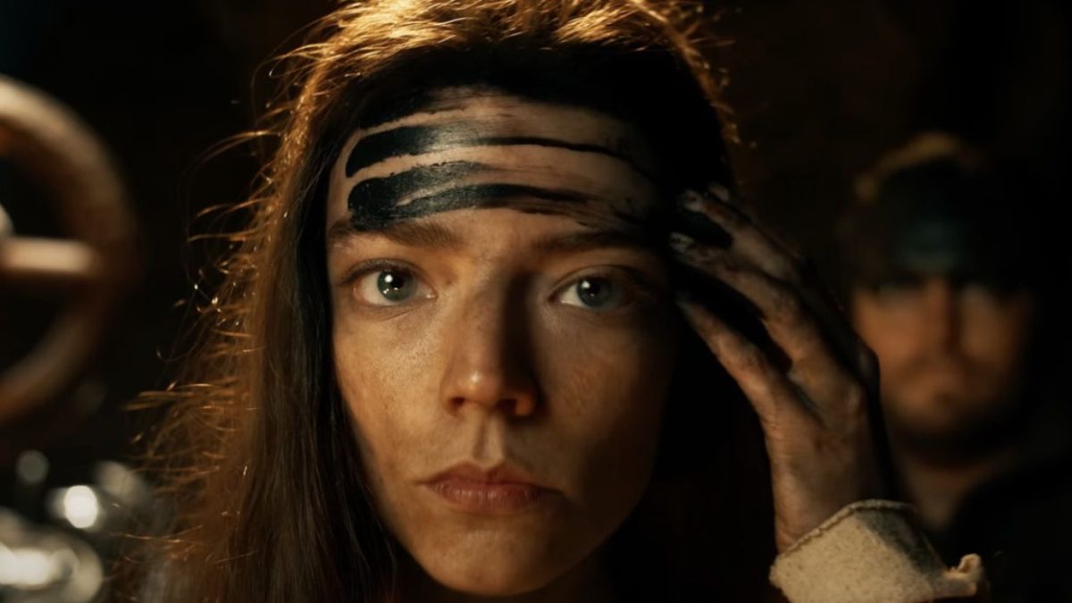 Furiosa (Anya Taylor-Joy) applies her war paint to her forehead while another soldier stoically stands by in the background in Furiosa